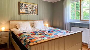 A bed with a colorful quilt, a nightstand, a wall painting, and a window with curtains.