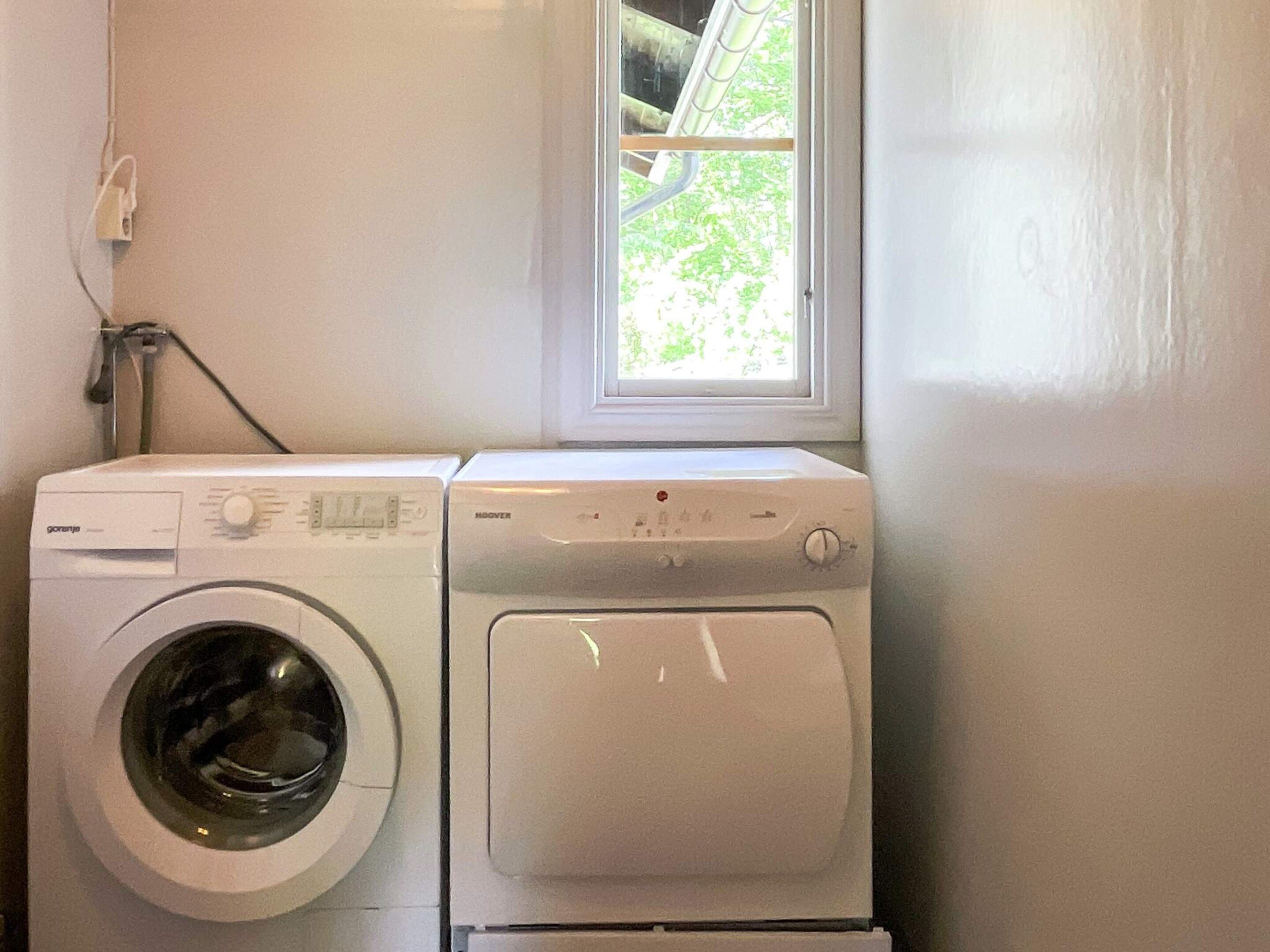 A laundry area with a front-loading washer and dryer, a window with a view of greenery, and a power outlet on the wall.