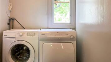 A laundry area with a front-loading washer and dryer, a window with a view of greenery, and a power outlet on the wall.