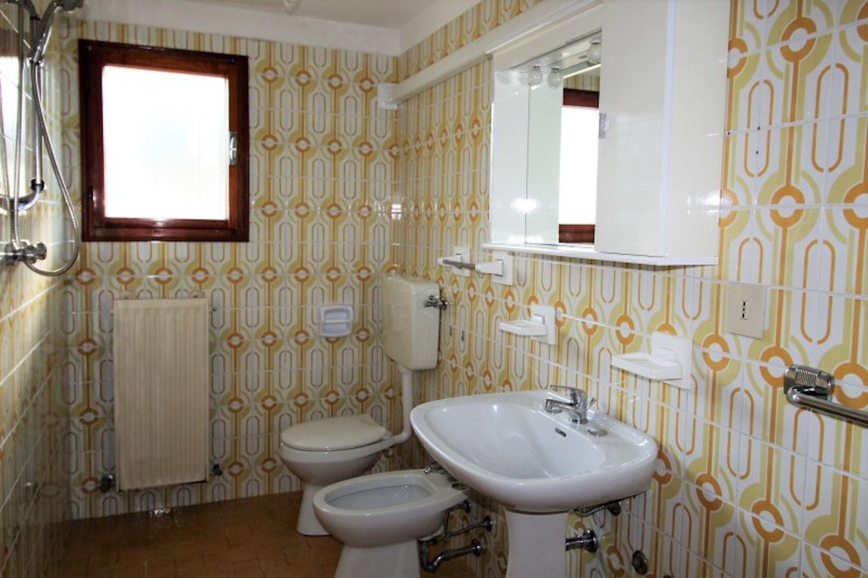 Casa de banho com sanita, bidé e lavatório, papel de parede estampado e janela.