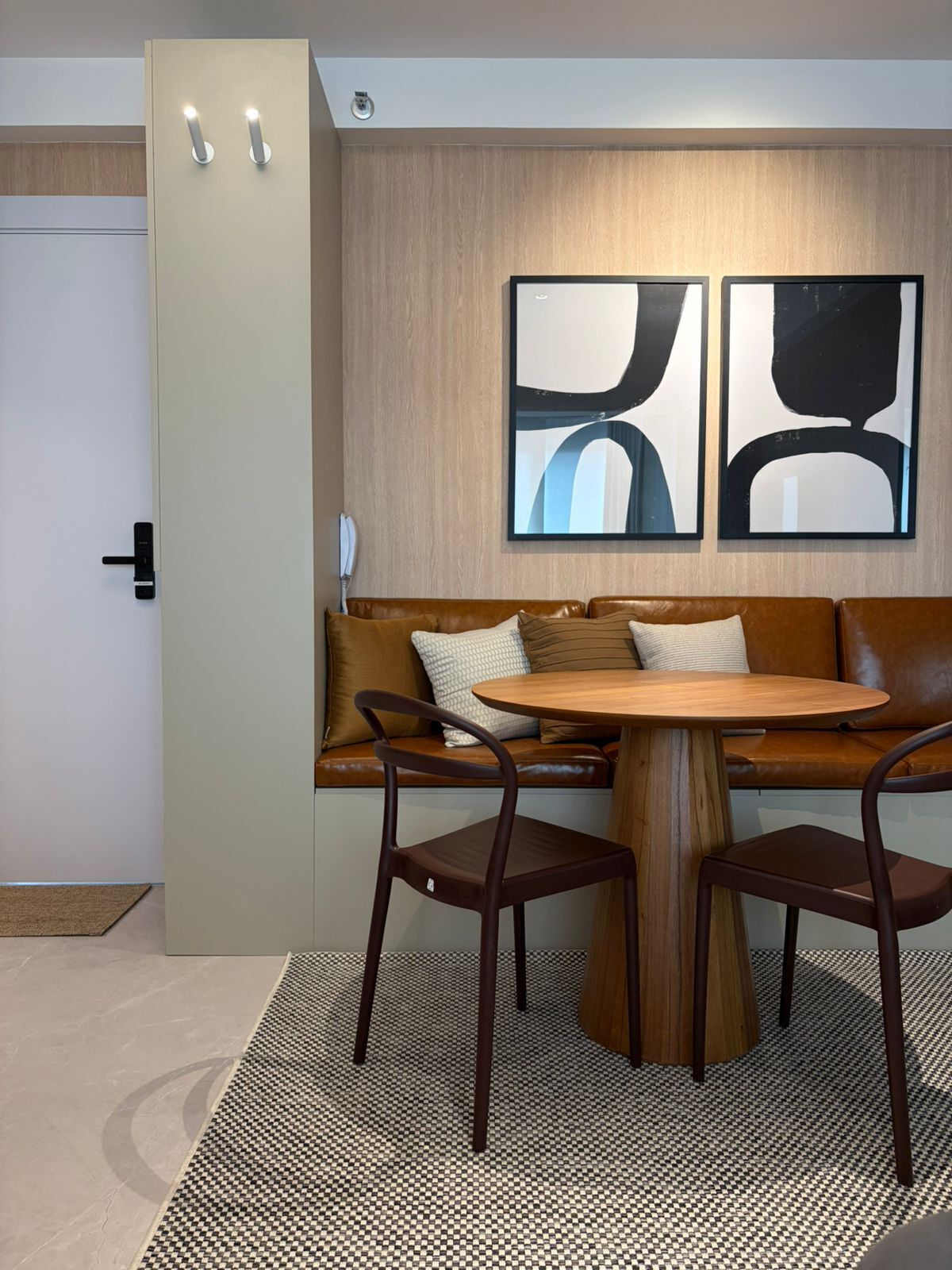 A modern interior with a brown leather sofa, two wooden chairs, a round wooden table, abstract wall art, and a light-colored door.