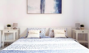 Ein Schlafzimmer mit einem Bett, zwei Nachttischen, zwei Kissen und einem Wandbild. Auf den Kissen ist der Schriftzug 'Maison' abgebildet.
