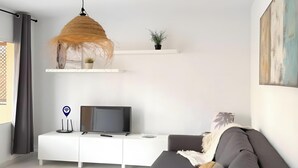 Ein modernes Wohnzimmer mit einem grauen Sofa, einem Fernsehgerät auf einem weißen Schrank, einer hängenden Leuchte und einem an der Wand befestigten Regal mit Pflanzen.