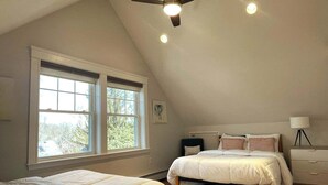 Ein Schlafzimmer mit großem Fenster, einem Bett, einem Nachttisch, einer Lampe und einem Deckenventilator.