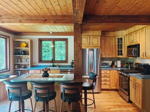 Eine Küche mit Holzausstattung, einem Edelstahlkühlschrank, einer Mikrowelle und einem Gasherd. Holzhocker umgeben eine zentrale Kochinsel.