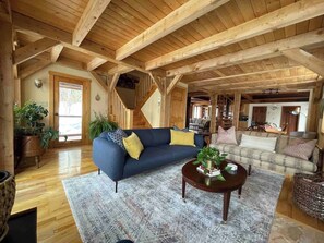 Ein geräumiges Wohnzimmer mit Holzbalken, einem blauen Sofa, einem runden Couchtisch und einer Treppe, die in eine weitere Ebene führt.