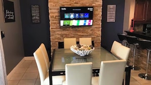A dining area with a glass table, white chairs, a stone wall, and a mounted TV displaying a menu. - 4 Bedroom Modern Pool home close to UCF (Oviedo)