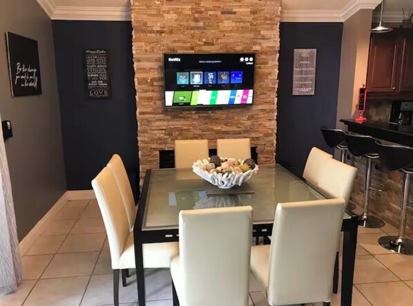 Un espace repas avec une table en verre, des chaises blanches, un mur en pierre et un téléviseur fixé au mur qui affiche un menu.