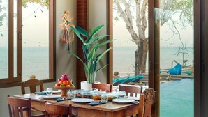 A dining table set with plates, glasses, and a fruit bowl, with a view of the sea through large windows. - Kampung Kecil Front Beach  (Kubu)