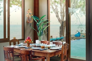 A dining table set with plates, glasses, and a fruit bowl, with a view of the sea through large windows.