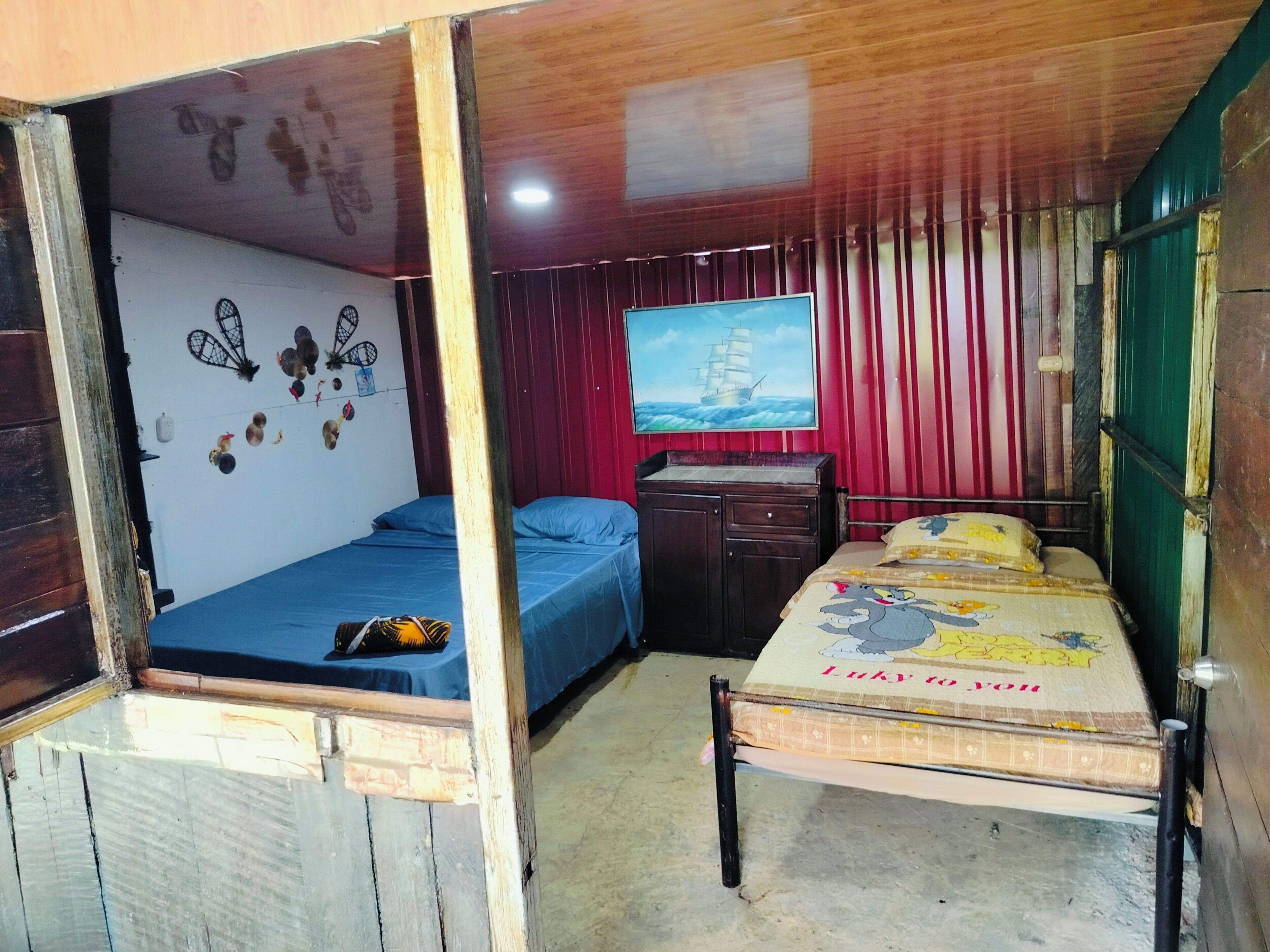 A room with two beds, a wooden ceiling, a red curtain, a wall-mounted TV, and a small cabinet.