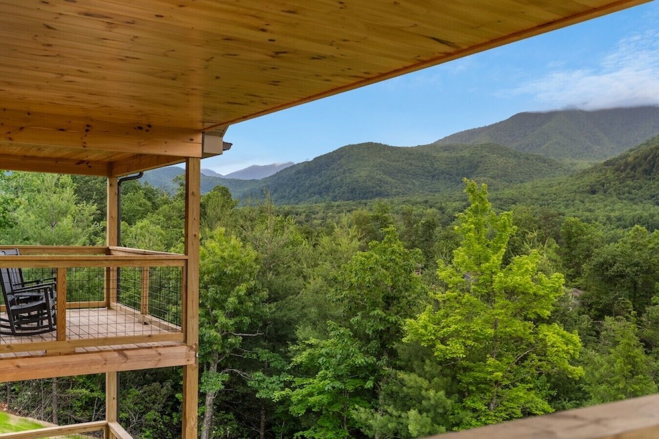 A wooden balcony with chairs, overlooking a forested valley with mountains in the background.