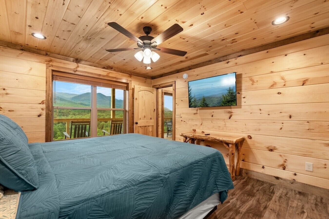 Dormitorio de cabaña de madera con cama, ventilador de techo, televisor y balcón con vistas a la montaña.