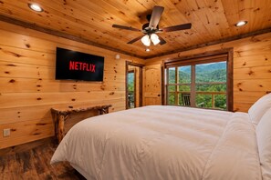 A wooden cabin bedroom with a bed, a ceiling fan, a TV mounted on the wall, and a window with a view of the outdoors. - Pool Cabin with 5 King Bedrooms (Cosby)