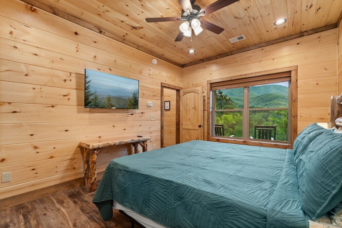 Un dormitorio de cabaña de madera con cama, ventilador de techo, televisión, ventana con vista a follaje, y un banco de madera.