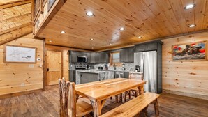 Interior de madera con mesa de comedor, bancos, zona de cocina y obras de arte en las paredes.