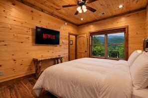 A wooden cabin bedroom with a bed, a television displaying 'NETFLIX,' a window with a view of mountains, and a ceiling fan. - Pool Cabin with 5 King Bedrooms (Cosby)