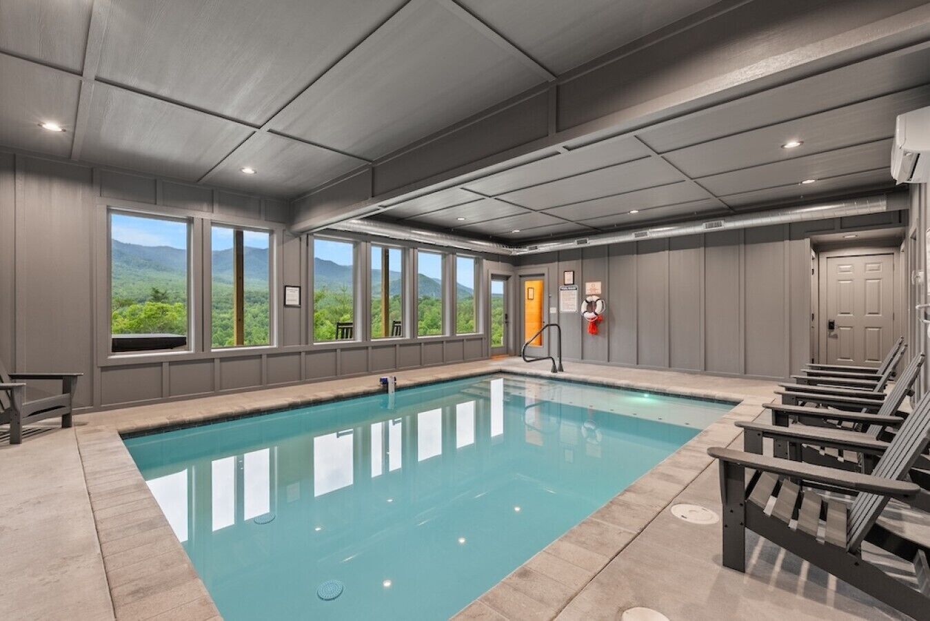 An indoor swimming pool with a view of greenery outside, a lifeguard chair, and a door with a sign.