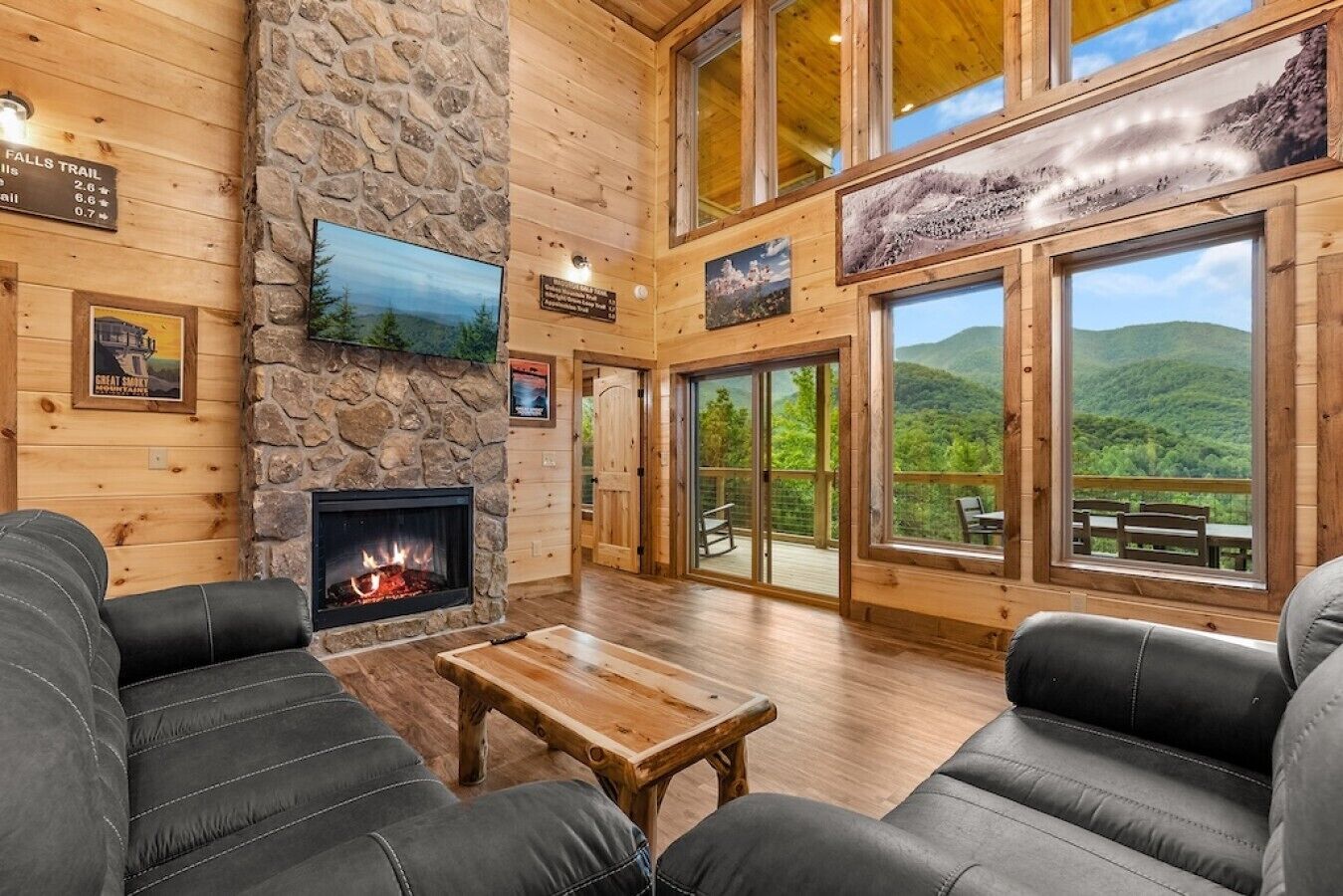 A log cabin interior with a fireplace, leather couches, a stone wall, and large windows offering mountain views.