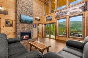 Ein rustikales Blockhaus-Interieur mit Kamin, Ledersofas, einer Steinwand und großen Fenstern mit Blick auf die Berge.