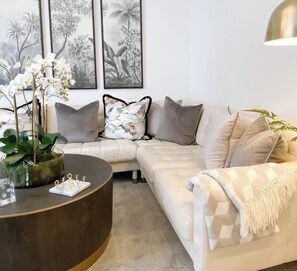 Een woonkamer met een beige bank, een ronde salontafel met een plant, en kunstwerken in lijsten aan de muur.