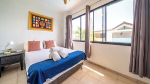 A hotel room with a bed, a nightstand, a ceiling fan, a window with curtains, and a painting on the wall. - Central One Bedroom Apartment on Third Floor (Sosúa)