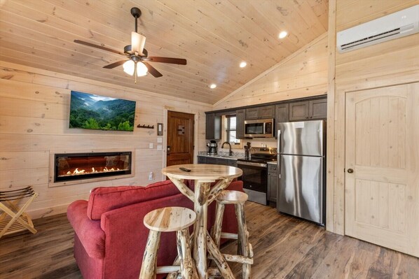 A cozy cabin interior with a fireplace, wooden walls, a kitchen with modern appliances, and a ceiling fan.