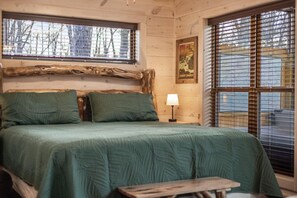 Un dormitorio de cabaña de madera con cama, almohadas, una mesita de noche, lámpara, banqueta y ventana con persianas.
