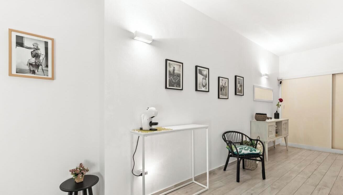 Une pièce avec un mur blanc, une petite table avec une lampe, une chaise et un meuble sur lequel se trouve un vase.