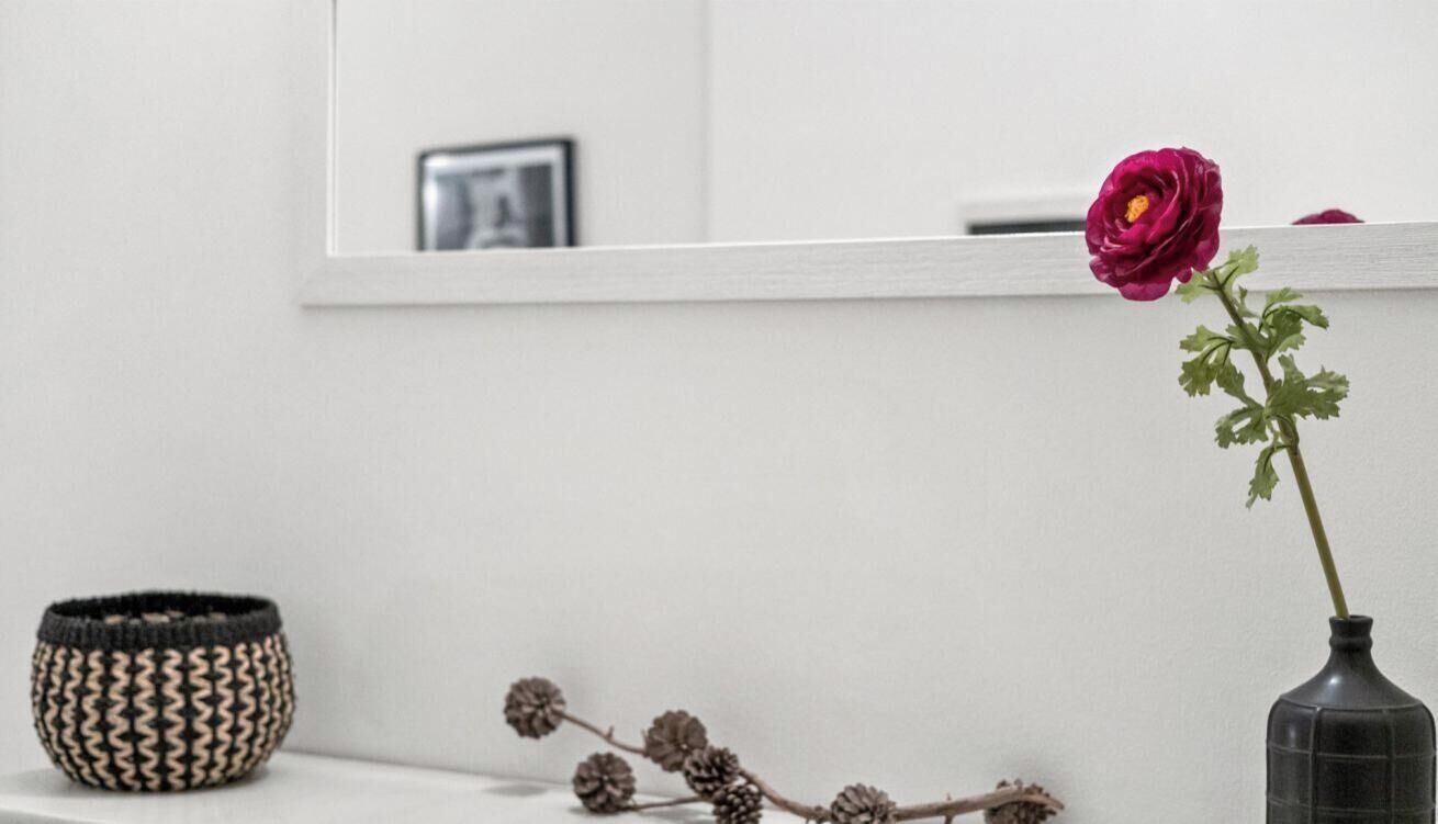Eine Vase mit einer einzelnen Blume, ein gewebter Korb und ein Zweig als Dekoration auf einer weißen Fläche.