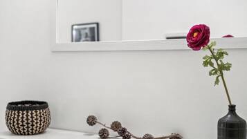 Eine Vase mit einer einzelnen Blume, ein gewebter Korb und ein Zweig als Dekoration auf einer weißen Fläche.