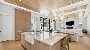 A modern kitchen with wooden cabinets, a large island, pendant lights, and a TV in the living area. - Retreat with Pool, LSV, Peloton and More (Santa Rosa Beach)