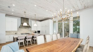 A modern kitchen with white cabinets, a large wooden dining table, and a chandelier. - Gulf Views with LSV, Pool, Peloton & More (Santa Rosa Beach)