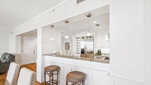 A modern kitchen with white cabinetry, a stainless steel refrigerator, and a bar area with stools. - Gulf View Condo with Community Amenities (Watersound)