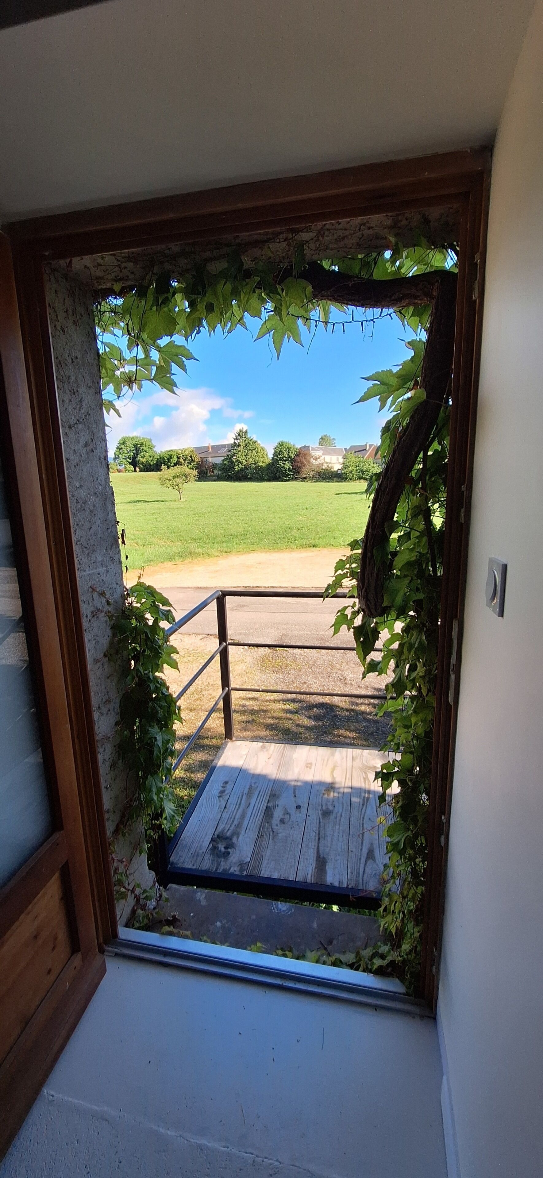 Une porte-fenêtre donnant sur une terrasse en bois, agréée d’une rambarde en métal et entourée de verdure.