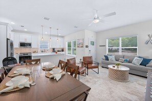 Eine moderne Küche mit weißen Schränken, ein Essbereich mit einem Holztisch für vier Personen und ein Wohnzimmer mit einem grauen Sofa und einem runden Couchtisch.