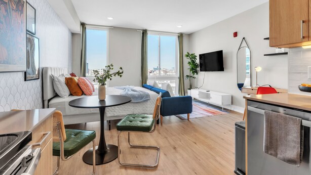 Quarto de hotel moderno e compacto, com cama, área de estar, cozinha compacta e janelas amplas com vista para a cidade.