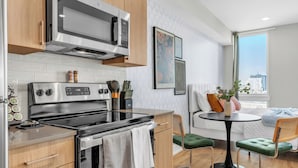 Cozinha moderna com micro-ondas, fogão e armários de madeira. Há uma área de refeições com mesa e cadeiras, e uma janela grande com vista para edifícios.