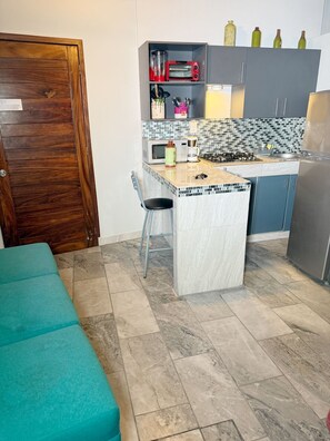 Eine kleine Küche mit Mikrowelle, Toaster und einer Kochinsel mit Spüle. Ein Stuhl und eine blaustürkise Bank stehen im Raum.