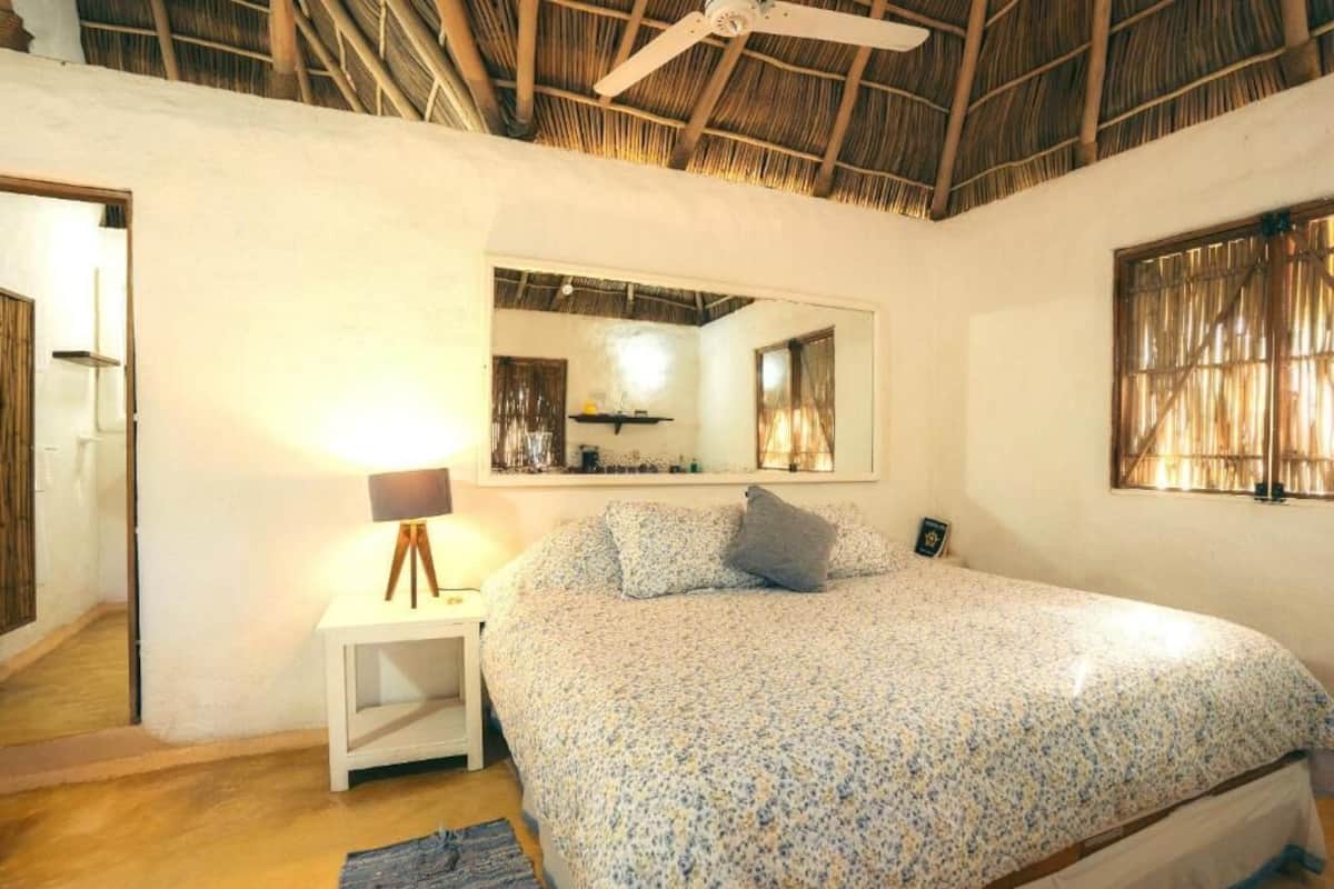 Quarto com cama, mesa de cabeceira com abajur, ventilador de teto, espelho e janela com cortinas.