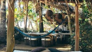 Una hamaca bajo una estructura de techo de paja, una cama con ropa de cama estampada y un entorno natural al aire libre.