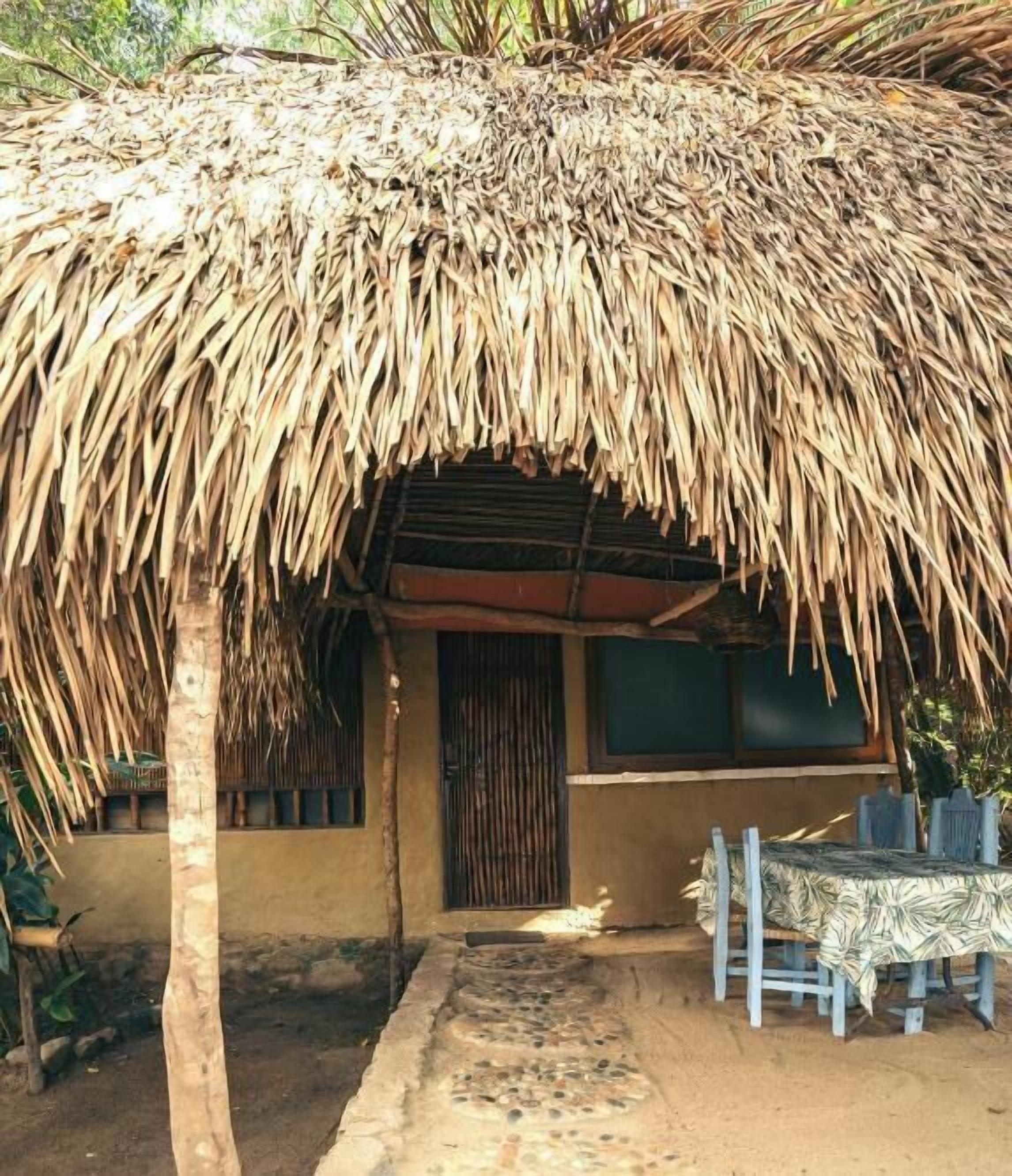 A thatched-roof hut with a door and windows, a table with a patterned tablecloth, and a chair.