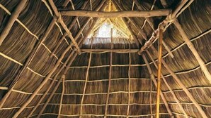 Una habitación con techo de paja, dos camas con ropa de cama azul, un aparador de madera y una ventana con cortinas.