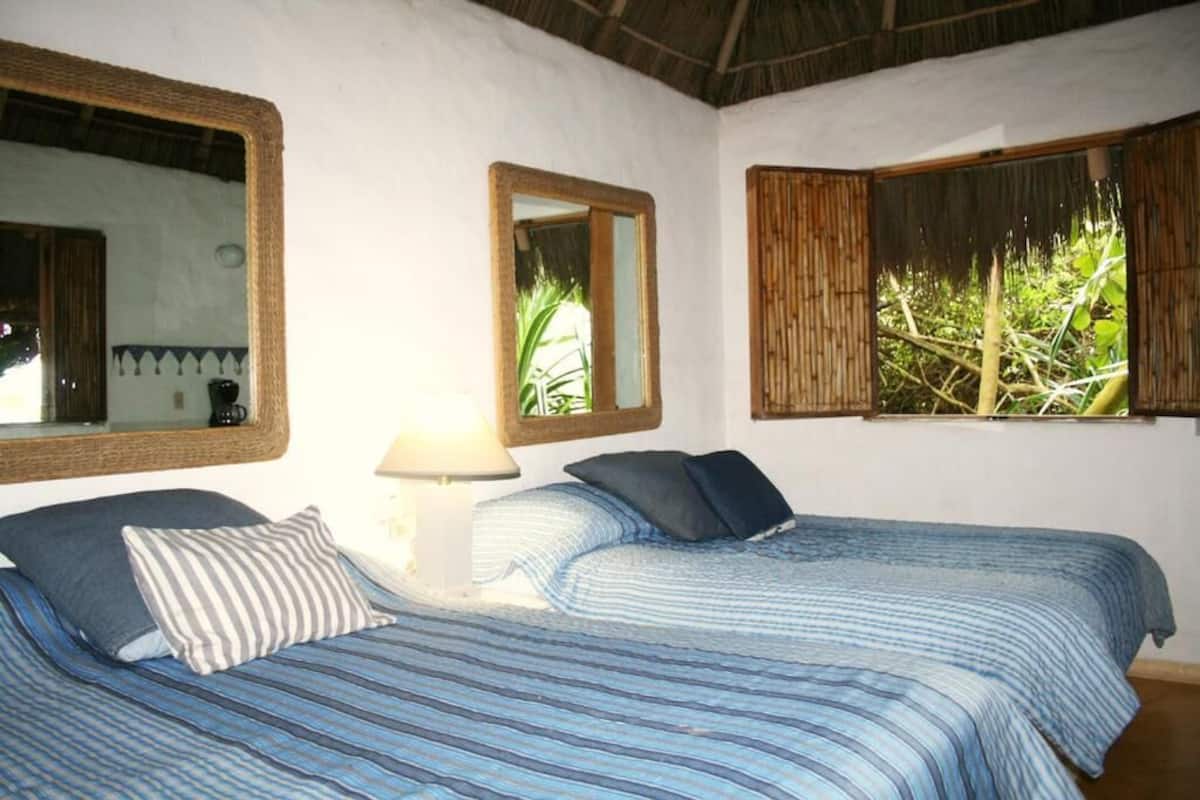 Quarto com duas camas, um espelho, uma luminária e janelas com vista para o verde.
