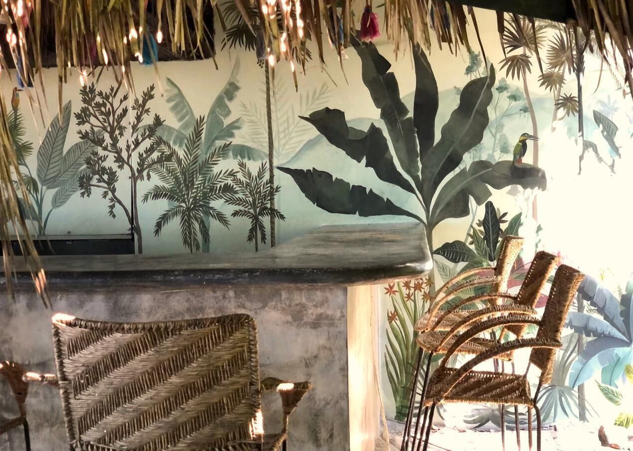 Una habitación con un mural de follaje tropical, muebles de mimbre y un techo de paja.