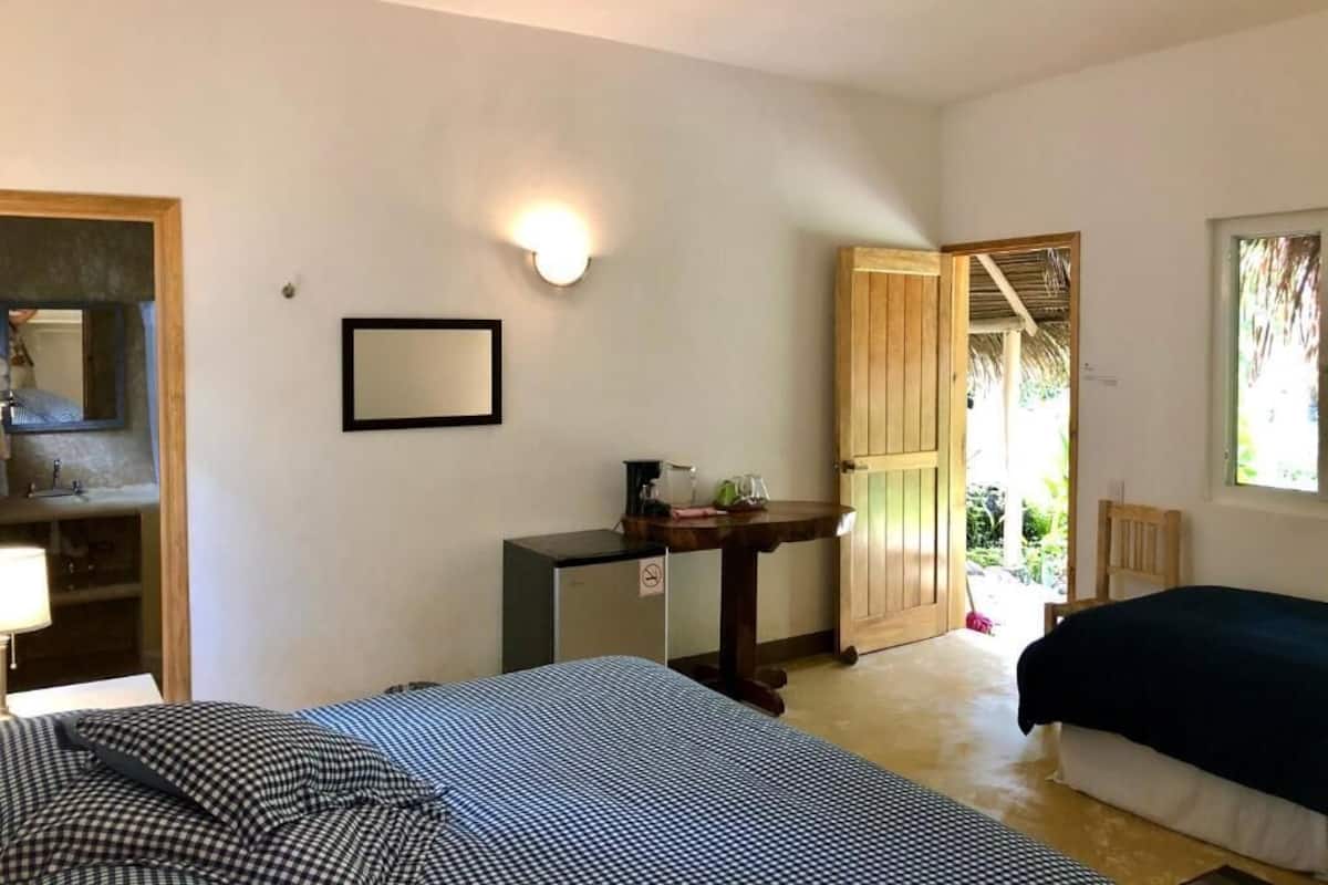 Quarto com cama, escrivaninha, espelho, luminária na parede e porta para área externa.