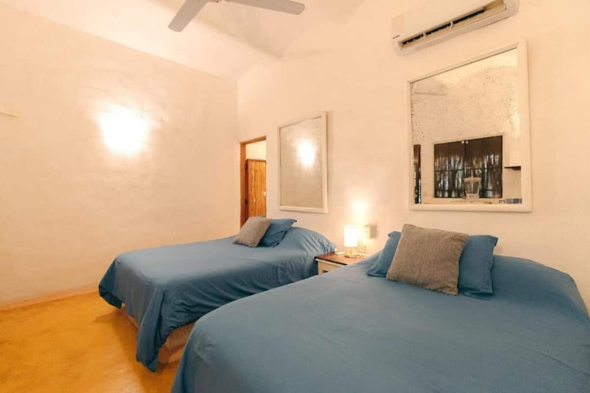 Quarto com duas camas, ventoinha no teto, ar condicionado, mesa de cabeceira com abajur e um quadro na parede.