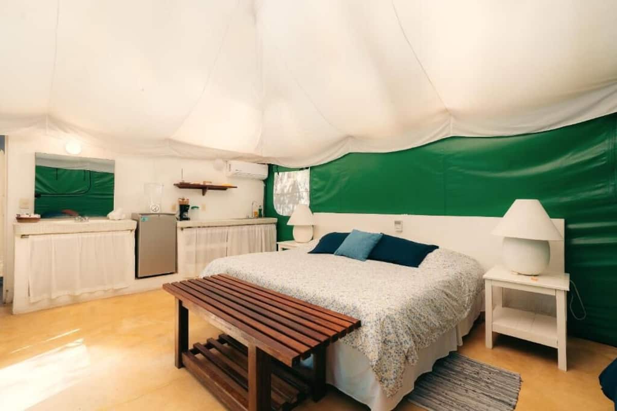 Um quarto com tenda, uma cama, mesa de cabeceira, luminária, banco e cozinha compacta.