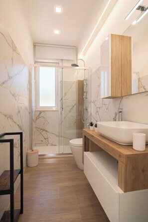 Ein modernes Badezimmer mit großem Spiegel, Waschtisch aus Holz, weißem Waschbecken und einer Duschkabine aus Glas.
