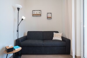 Ein modernes Wohnzimmer mit einem schwarzen Sofa, einem kleinen Beistelltisch mit Taschentuchbox und Buch, einer Stehlampe und zwei Wandregalen mit Pflanzen.